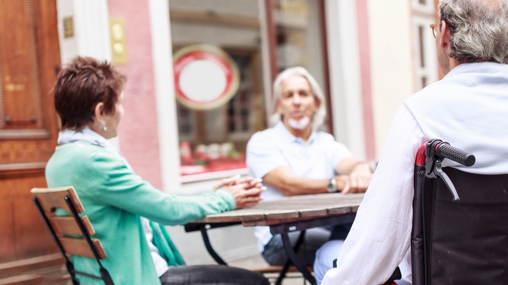 Menschen sitzen vor einem Café, ein Mann sitzt im Rollstuhl