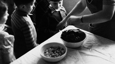 Kinder sehen ihrer Mutter zu wie sie zwei Schüsseln mit Essen vorbereitet um 1945.
