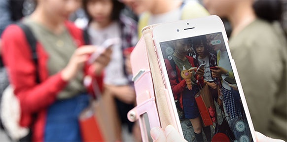 Eine Hand hält ein Handy mit rosa Hülle, am Display sieht man eine Gruppe japanischer Mädchen und ein Pokemon.