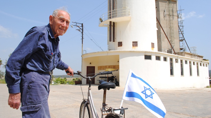 Chaim Miller im blauen Arbeiteranzug mit seinem Fahrrad, darauf ist eine Israel-Fahne befestigt.