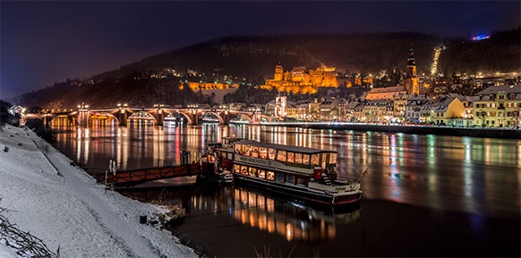 Heidelberg in einer Winternacht: ein Schiff fährt am Fluss, auf dem sich die bunten Lichter der Stadt spiegeln.