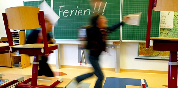 Tafel mit Aufschrift "Ferien!", davor Kinder mit ihren Zeugnissen