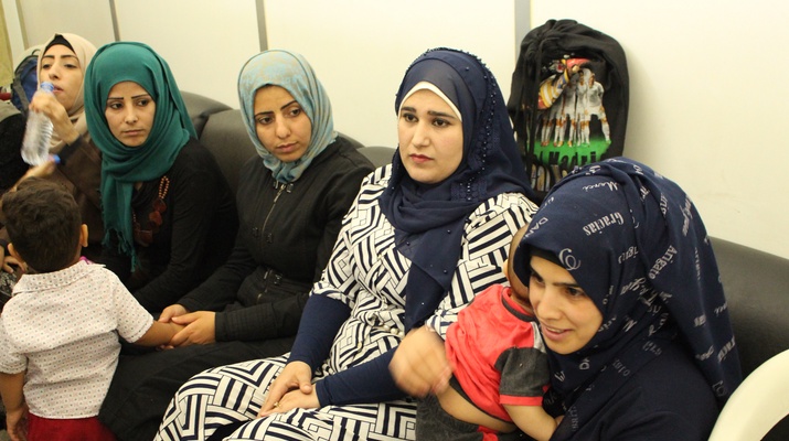 Eine Gruppe von Frauen mit färbigen Kopftüchern sitzt auf einer Bank in einem Gesundeitszentrum. Manche haben Kinder am Schoß.