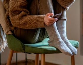 Junge Frau hockt mit angezogenen Beinen auf einem Sessel, in der Hand ein Smartphone