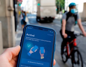 Mann hält in Frankreich ein Smartphone in Händen, in Hintergrundunschärfe ein Radfahrer mit Mundschutz