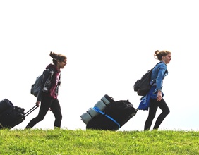 Zwei Mädchen mit ihren Koffern unterwegs auf einer Wiese.