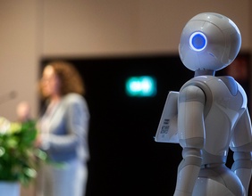 Ein weißer Roboter steht auf der Bühne der Konferenz. Im Hintergrund verschwommen die Referentin.