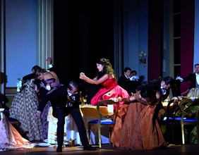 Szene aus "Teseo", Frau in einem Roten Kleid umgeben von anderen DarstellerInnen