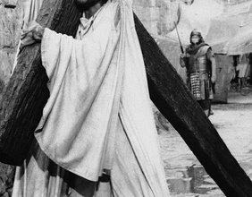 Mann als Jesus verkleidet trägt ein Kreuz, dahinter ein Römer