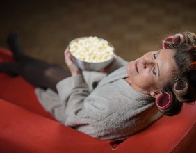 Eine Frau im Bademantel und mit Lockenwicklern auf einem Sofa liegend und Popcorn essend