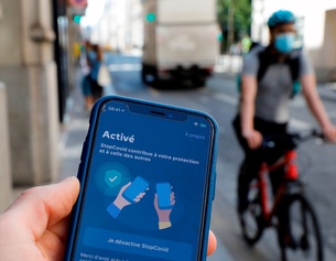 Mann hält in Frankreich ein Smartphone in Händen, in Hintergrundunschärfe ein Radfahrer mit Mundschutz