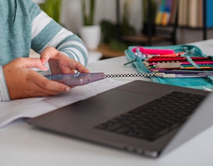 Kind hält Smartphone in Händen und sitzt an einem Tisch mit Laptop und Stiften