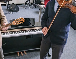Zwei ältere Herren spielen ein Instrument in einem Klassenzimmer/Musikschule: Laute und Violine, dahinter ein Keyboard