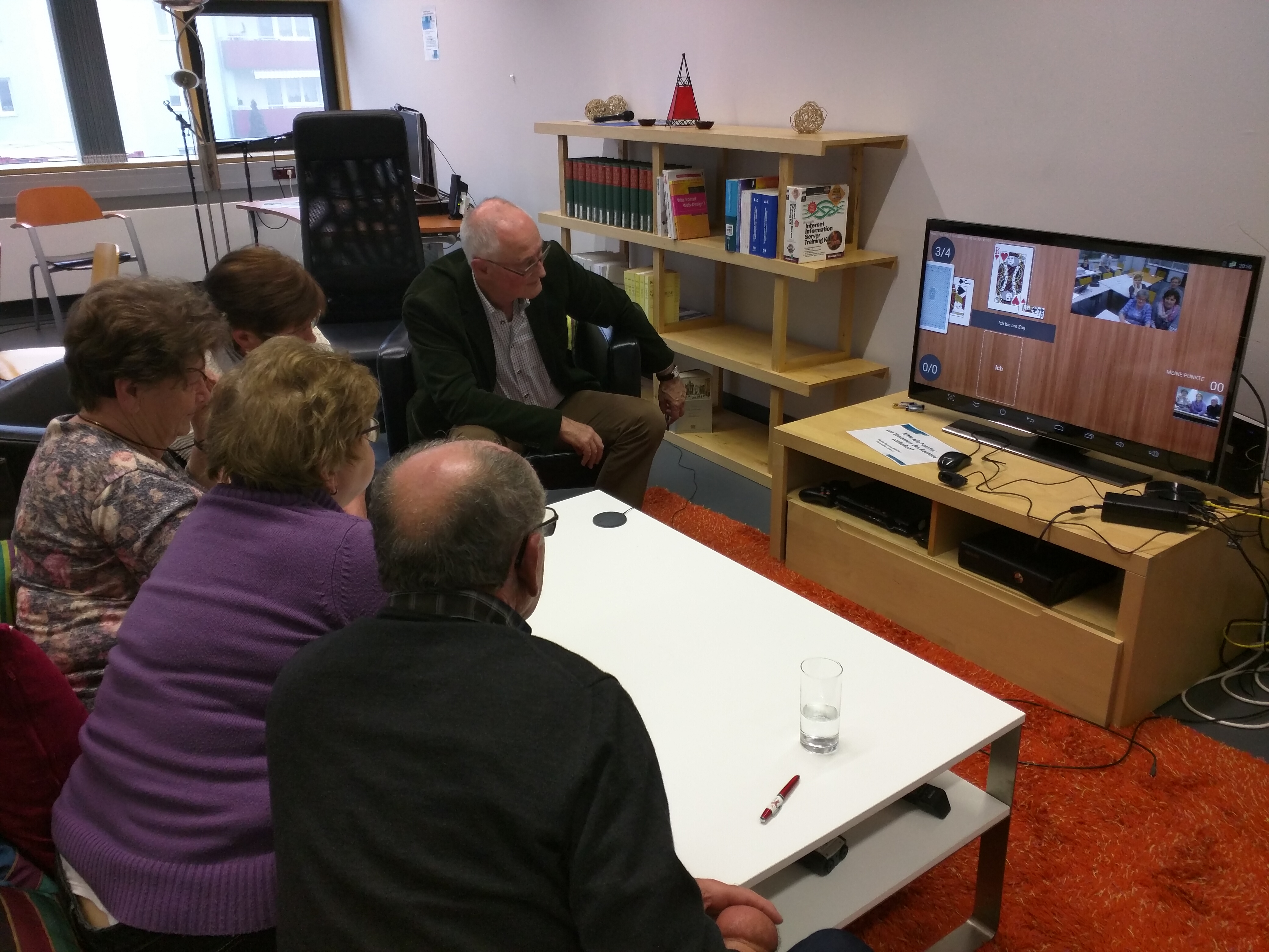 eine Gruppe Senior:innen testet Umbrello. Fünf Personen sitzen vor einem Fernseher auf dem eine andere Gruppe von Senir:innen und ein Schnapskarten-Spiel zu sehen sind