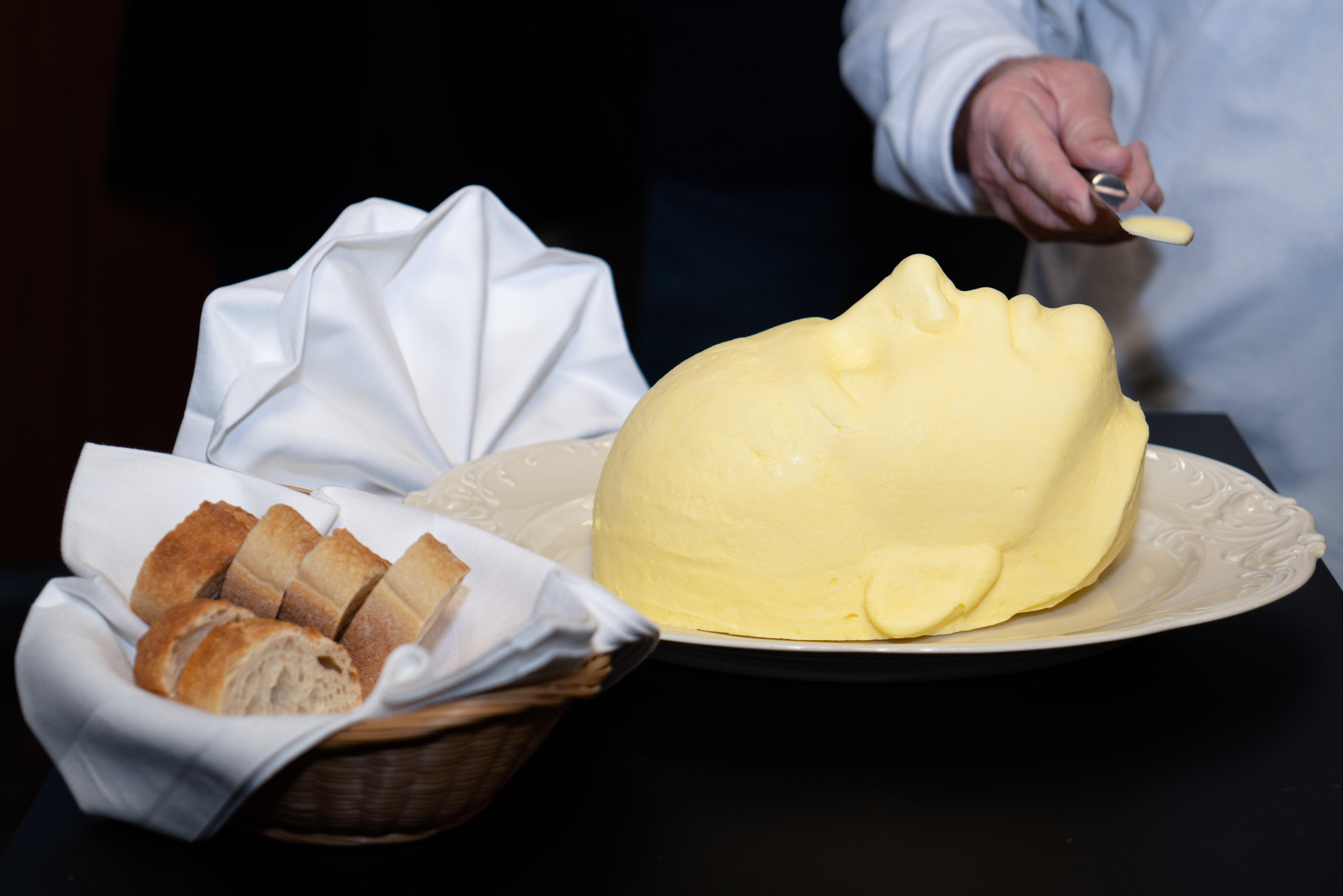 Selbstportrait der Künstlerin aus Butter; links daneben ein Korb mit Brot; rechts eine Hand mit einer Messerspitze Butter;